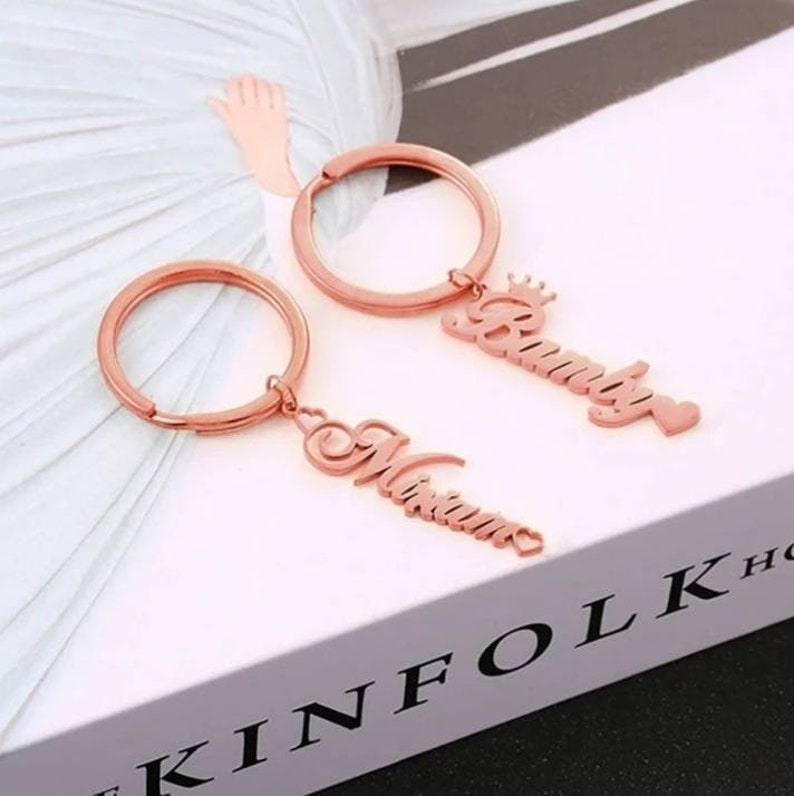 May include: Two rose gold keychains with the names "Brittany" and "Miriam" engraved on them. The keychain with the name "Brittany" has a small crown above the name. Both keychains have a small heart charm at the bottom.