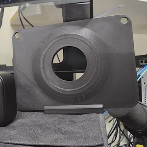 May include: A black dashcam with a circular lens and a matching black mounting bracket. The lens has a colorful, reflective surface. The bracket has a large circular opening. The items are on a gray surface.