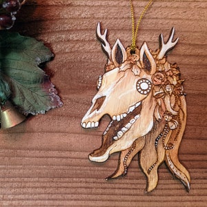 May include: A wooden ornament shaped like a deer skull with antlers, decorated with flowers and intricate details. The ornament is painted in shades of brown and white, and has a gold string attached for hanging.