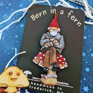 May include: Wooden ornament of a gnome wearing a blue jacket and a face mask, sitting on a red and white mushroom. The ornament is labeled "Bern in a fern handmade in Frederick, MD".