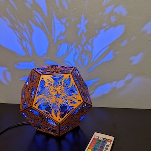 Puede incluir: Una l&aacute;mpara geom&eacute;trica de madera con un dise&ntilde;o intrincado proyecta una luz azul sobre una pared, creando una sombra de una flor. La l&aacute;mpara est&aacute; conectada a un mando a distancia con opciones de color.