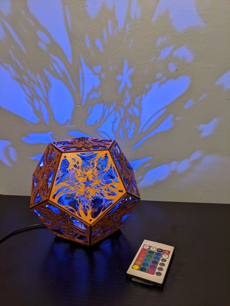 Puede incluir: Una l&aacute;mpara geom&eacute;trica de madera con un dise&ntilde;o intrincado proyecta una luz azul sobre una pared, creando una sombra de una flor. La l&aacute;mpara est&aacute; conectada a un mando a distancia con opciones de color.