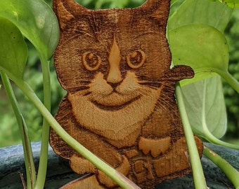 May include: A wooden cat plant stake with a smiling face and a small, pointed stake for insertion into soil. The cat is brown with white markings and is surrounded by green leaves. The cat is a decorative garden accessory.