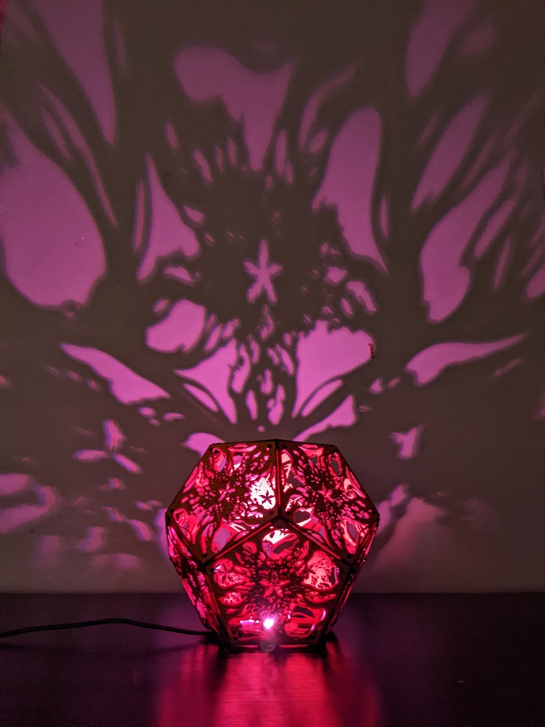 Puede incluir: Una luz rosa proyecta intrincadas sombras florales en la pared detr&aacute;s de una l&aacute;mpara roja geom&eacute;trica con un delicado dise&ntilde;o de corte l&aacute;ser.