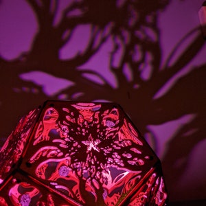 Puede incluir: Una pantalla de l&aacute;mpara de madera roja con un dise&ntilde;o geom&eacute;trico. La pantalla est&aacute; iluminada desde el interior, proyectando un brillo rosado.