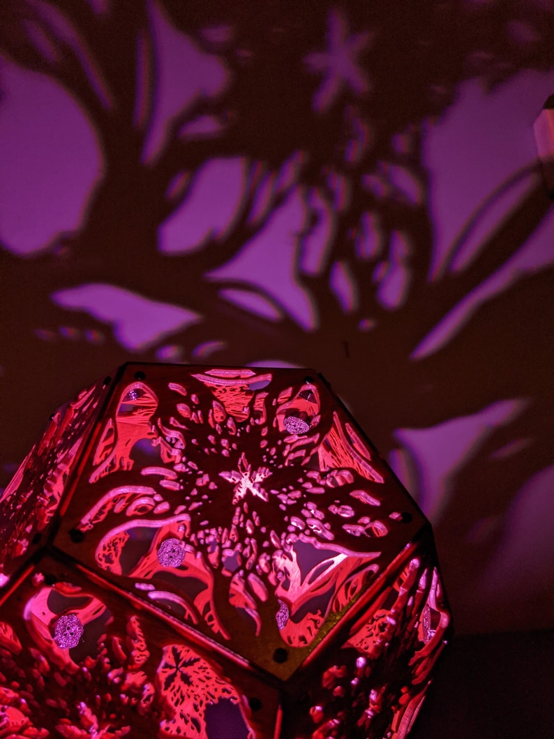 Puede incluir: Una pantalla de l&aacute;mpara de madera roja con un dise&ntilde;o geom&eacute;trico. La pantalla est&aacute; iluminada desde el interior, proyectando un brillo rosado.