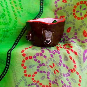 May include: A small brown ceramic planter with a pink rim and two white hearts painted on the front. The planter is sitting on a green fabric with a red and purple pattern.