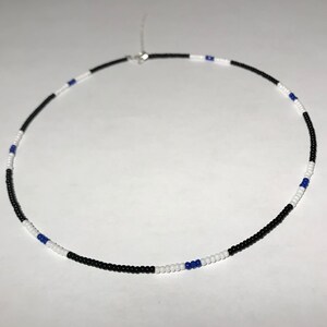 May include: A beaded necklace featuring a pattern of black, white, and blue beads. The necklace is composed of small, round beads strung together, with a silver-colored clasp. The design alternates colors, creating a visually appealing accessory.