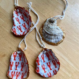 May include: Three decorative oyster shell ornaments featuring a white interior with a repeating red lobster pattern, gold-painted edges, and a white string loop with a small white bead for hanging. One shell shows the textured, natural gray exterior.