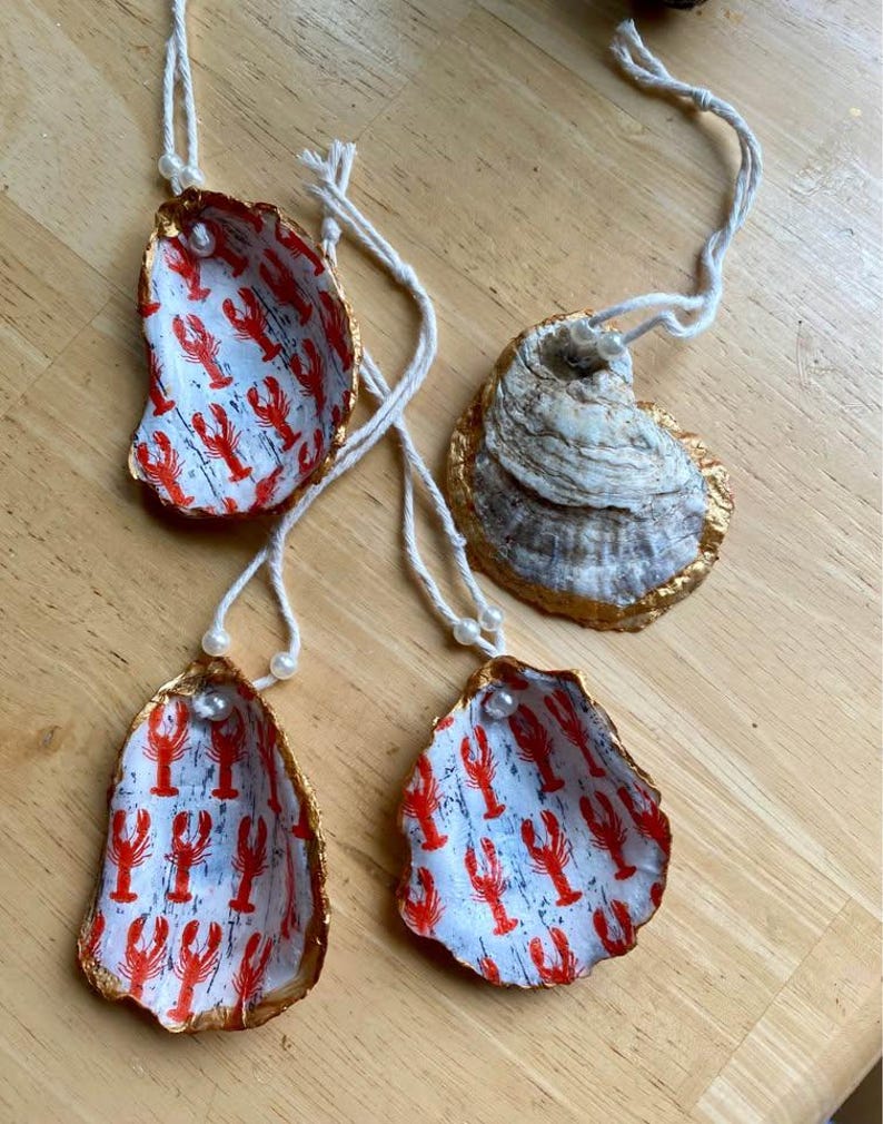 May include: Three decorative oyster shell ornaments featuring a white interior with a repeating red lobster pattern, gold-painted edges, and a white string loop with a small white bead for hanging. One shell shows the textured, natural gray exterior.