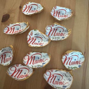 May include: A collection of nine oyster shells, each featuring a gold-painted edge and a decorative interior design. The interior of each shell displays the word Maine written in red cursive script, accompanied by small blue stars and illustrations.