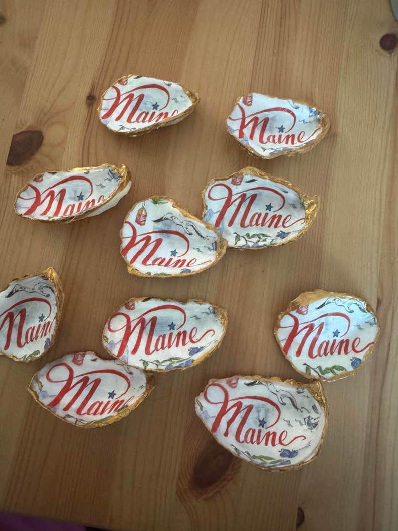 May include: A collection of nine oyster shells, each featuring a gold-painted edge and a decorative interior design. The interior of each shell displays the word Maine written in red cursive script, accompanied by small blue stars and illustrations.