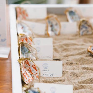 May include: A white rectangular tray filled with sand containing multiple oyster shells with gold-painted edges. Each shell holds a white rectangular place card featuring a blue seashell icon, the text TABLE 4, and the name ANDREY.