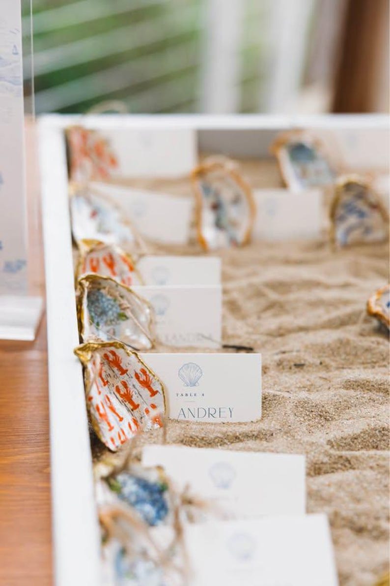 May include: A white rectangular tray filled with sand containing multiple oyster shells with gold-painted edges. Each shell holds a white rectangular place card featuring a blue seashell icon, the text TABLE 4, and the name ANDREY.