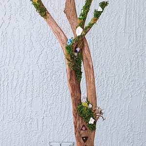 Könnte beinhalten: Ein Treibholzbaum, verziert mit Moos, weißen Blüten und Kristallen, in einem blau-weißen Keramiktopf auf einem schwarzen Metallständer. Das Treibholz hat ein geschnitztes Design mit sieben Chakra-Steinen.