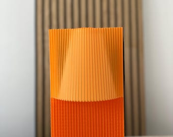 Puede incluir: Un objeto decorativo rectangular de dos tonos. La mitad superior es de color naranja claro y la mitad inferior es de color naranja más oscuro. El objeto tiene una textura acanalada, con líneas verticales. El fondo presenta una pared de listones de madera.