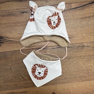 May include: A white hat with two top knots and ear flaps, paired with a matching triangular bib. Both items feature a brown and white lion face print and a tan interior lining. The hat includes braided chin straps and the bib has metal snaps.