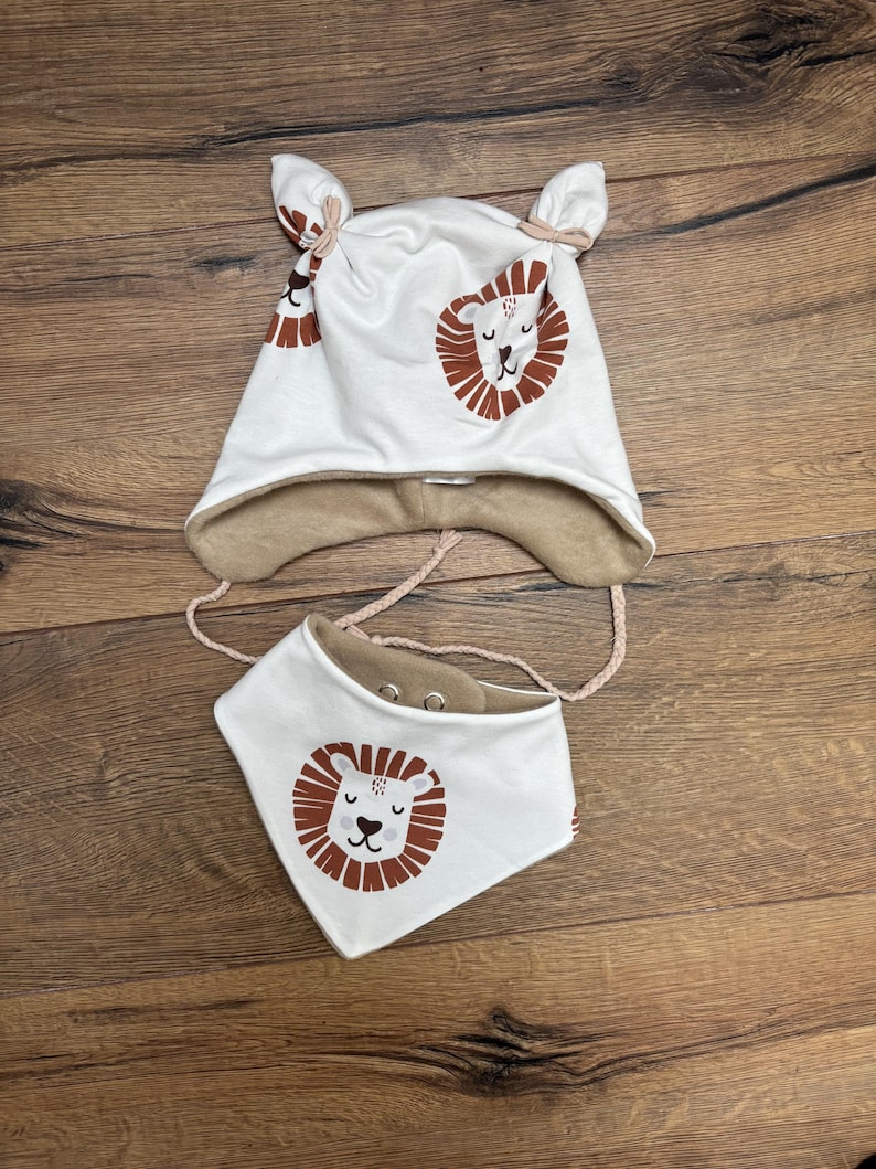 May include: A white hat with two top knots and ear flaps, paired with a matching triangular bib. Both items feature a brown and white lion face print and a tan interior lining. The hat includes braided chin straps and the bib has metal snaps.