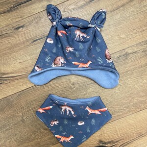 May include: A dark blue beanie with two knotted top corners and a matching triangular bandana bib. Both items feature a repeating pattern of orange foxes, deer, and small hedgehogs interspersed with green pine trees on a dark blue background.