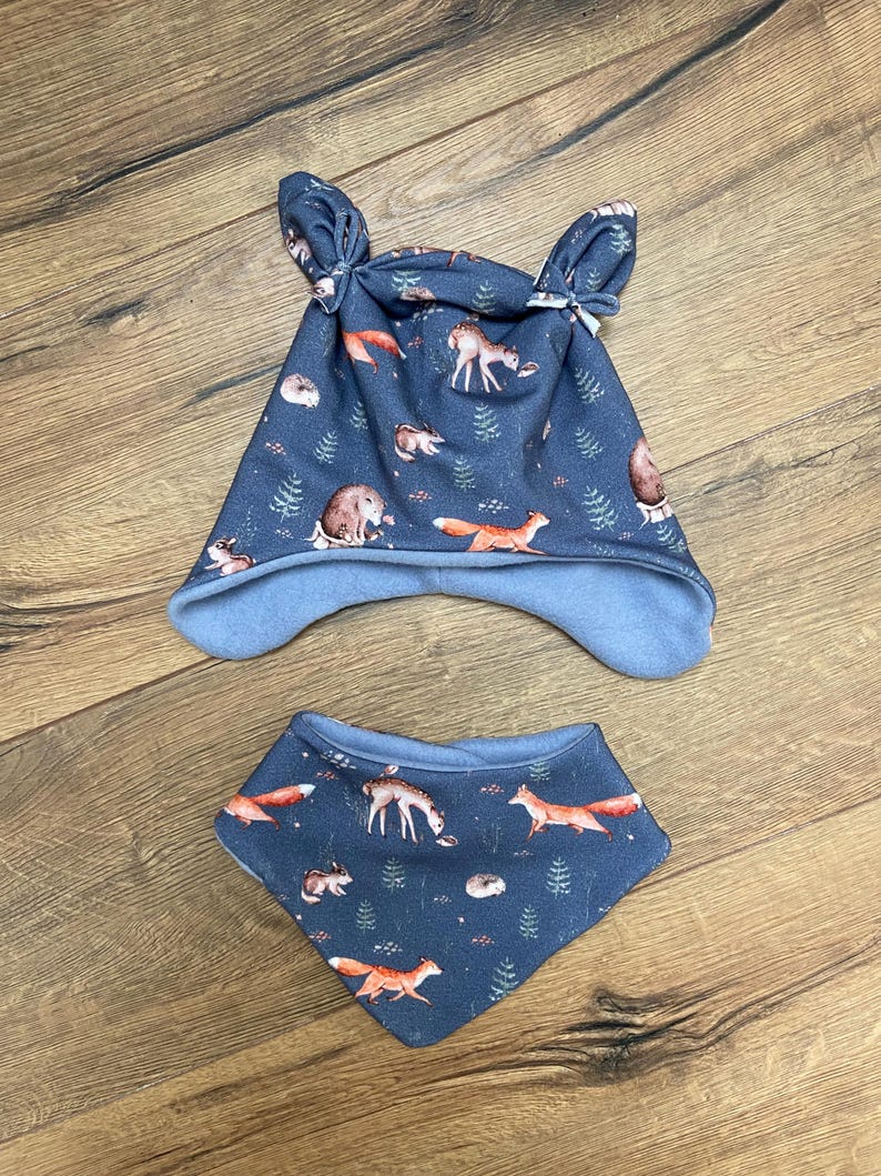 May include: A dark blue beanie with two knotted top corners and a matching triangular bandana bib. Both items feature a repeating pattern of orange foxes, deer, and small hedgehogs interspersed with green pine trees on a dark blue background.