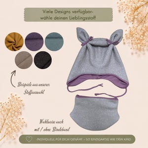 Könnte beinhalten: Eine graue Mütze mit zwei spitzen Ohren und passendem Schlauchschal. Die Mütze hat ein violettes Innenfutter und geflochtene Bindebänder. Fünf kreisförmige Stoffmuster in Senfgelb, Violett, Petrol, Beige und Schwarz sind abgebildet.