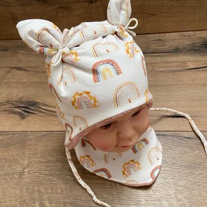 May include: A white beanie with two knotted top corners and a matching neck warmer, both featuring a repeating pattern of rainbows in shades of orange, yellow, and brown. The set includes braided white ties and has a soft pink inner lining.