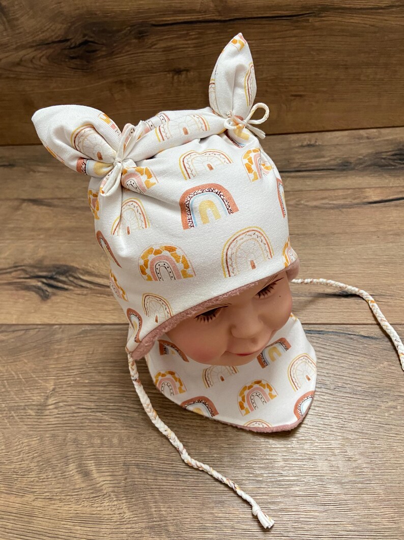 May include: A white beanie with two knotted top corners and a matching neck warmer, both featuring a repeating pattern of rainbows in shades of orange, yellow, and brown. The set includes braided white ties and has a soft pink inner lining.