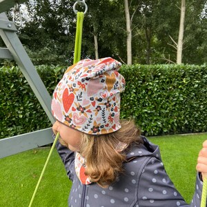 May include: A beanie hat featuring a white background with a repeating pattern of red and pink hearts, black flowers, and green leaves. The hat is worn with a matching neck warmer, both made of fabric with a soft, smooth texture and vibrant colors.