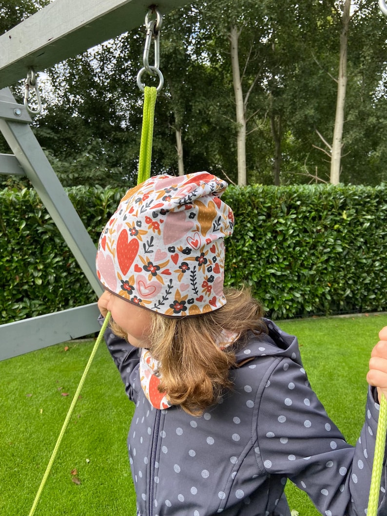 May include: A beanie hat featuring a white background with a repeating pattern of red and pink hearts, black flowers, and green leaves. The hat is worn with a matching neck warmer, both made of fabric with a soft, smooth texture and vibrant colors.