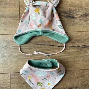 May include: A light pink hat with two top knots, ear flaps, and braided ties, featuring a pattern of rabbits, carrots, and flowers. The hat has a solid sage green lining. Included is a matching triangular bandana bib with two snap buttons.