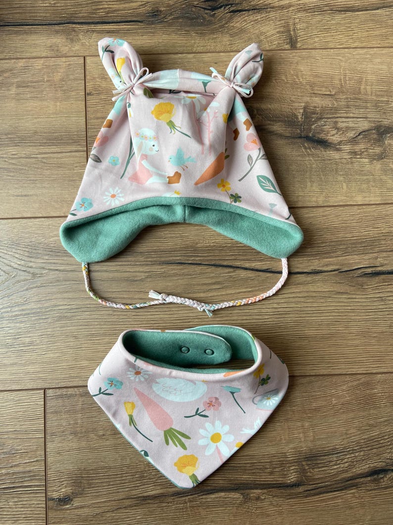 May include: A light pink hat with two top knots, ear flaps, and braided ties, featuring a pattern of rabbits, carrots, and flowers. The hat has a solid sage green lining. Included is a matching triangular bandana bib with two snap buttons.