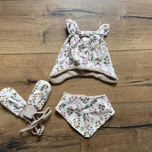 May include: A set consisting of a hat with two small ears on top, a pair of mittens with attached braided strings, and a triangular bandana bib. All items feature a white fabric base with a repeating pattern of brown and purple leaves.
