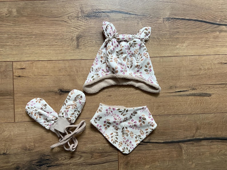 May include: A set consisting of a hat with two small ears on top, a pair of mittens with attached braided strings, and a triangular bandana bib. All items feature a white fabric base with a repeating pattern of brown and purple leaves.