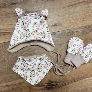 K&ouml;nnte beinhalten: Ein Set bestehend aus einer M&uuml;tze mit zwei kleinen Ohren, einem dreieckigen Halstuch und einem Paar F&auml;ustlinge. Alle Teile bestehen aus wei&szlig;em Stoff mit einem Muster aus braunen und rosafarbenen Bl&auml;ttern und Zweigen, gef&uuml;ttert mit beigem Stoff.