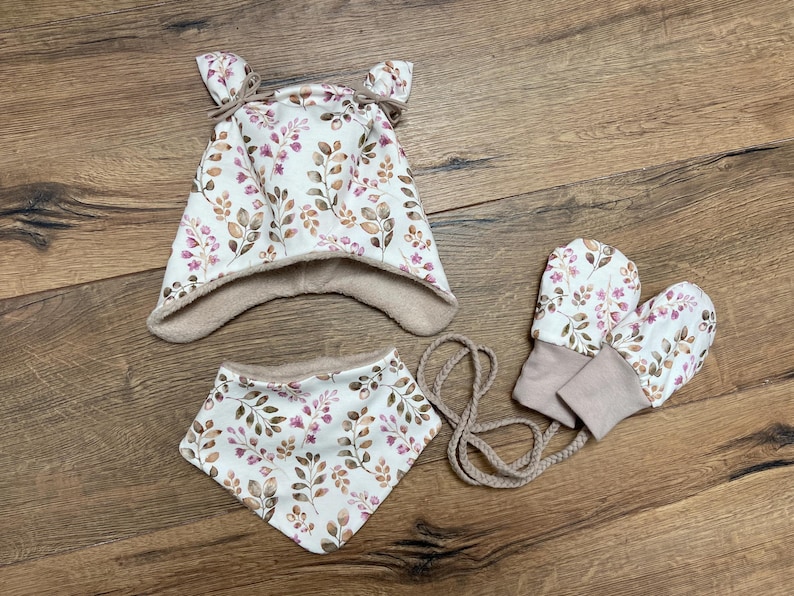K&ouml;nnte beinhalten: Ein Set bestehend aus einer M&uuml;tze mit zwei kleinen Ohren, einem dreieckigen Halstuch und einem Paar F&auml;ustlinge. Alle Teile bestehen aus wei&szlig;em Stoff mit einem Muster aus braunen und rosafarbenen Bl&auml;ttern und Zweigen, gef&uuml;ttert mit beigem Stoff.