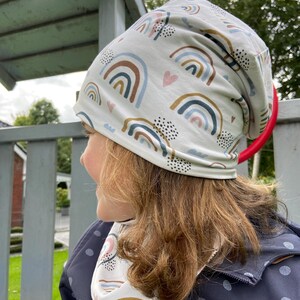 May include: A slouchy beanie hat and matching neck warmer featuring a repeating pattern of rainbows, small pink hearts, and black dotted clusters on a light cream background. The fabric has a soft, smooth texture and a relaxed, comfortable fit.