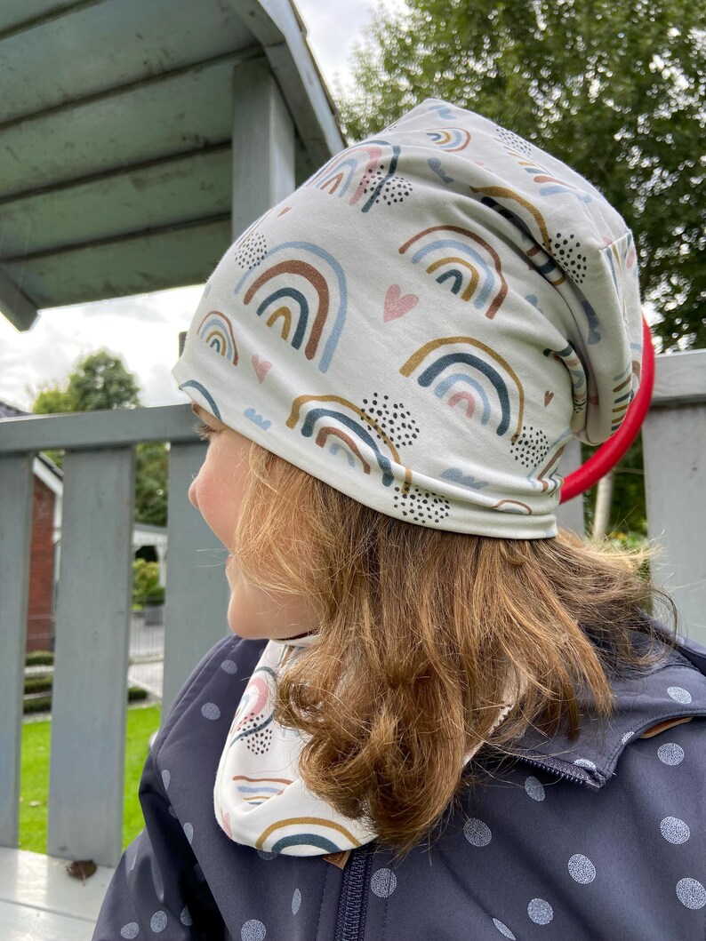 May include: A slouchy beanie hat and matching neck warmer featuring a repeating pattern of rainbows, small pink hearts, and black dotted clusters on a light cream background. The fabric has a soft, smooth texture and a relaxed, comfortable fit.