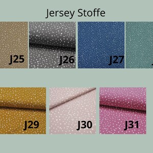 May include: A collection of seven jersey fabric swatches labeled J25 through J31, each featuring a white speckled pattern on solid backgrounds of tan, gray, blue, teal, mustard yellow, light pink, and magenta. The text Jersey Stoffe is visible.