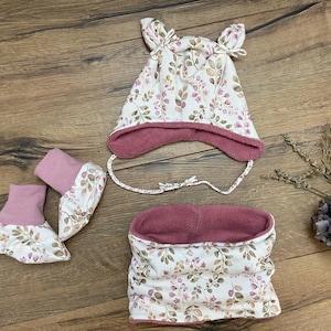 K&ouml;nnte beinhalten: Ein Set bestehend aus einer M&uuml;tze mit Ohrenklappen und Bindeb&auml;ndern, einem Paar Sch&uuml;hchen und einem Schlauchschal. Alle Teile sind aus cremefarbenem Stoff mit einem botanischen Druck aus braunen und rosa Bl&auml;ttern und altrosa gef&uuml;ttert.