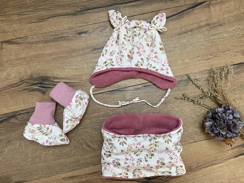 K&ouml;nnte beinhalten: Ein Set bestehend aus einer M&uuml;tze mit Ohrenklappen und Bindeb&auml;ndern, einem Paar Sch&uuml;hchen und einem Schlauchschal. Alle Teile sind aus cremefarbenem Stoff mit einem botanischen Druck aus braunen und rosa Bl&auml;ttern und altrosa gef&uuml;ttert.