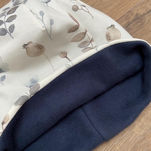 May include: A beanie hat featuring an off-white exterior with a printed pattern of muted brown and gray botanical elements, including leaves and seed pods. The interior of the hat is lined with a solid dark navy blue fleece fabric.