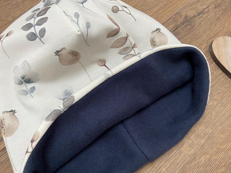 May include: A beanie hat featuring an off-white exterior with a printed pattern of muted brown and gray botanical elements, including leaves and seed pods. The interior of the hat is lined with a solid dark navy blue fleece fabric.