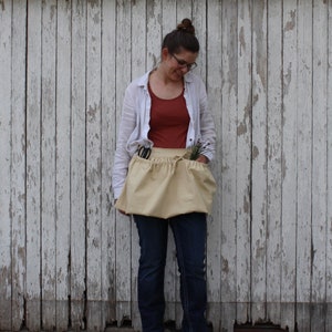 May include: A light tan gardening apron with two large pockets, holding scissors and small plants. The apron is tied at the waist and worn over a red top and blue jeans. The background is a weathered white wooden wall.