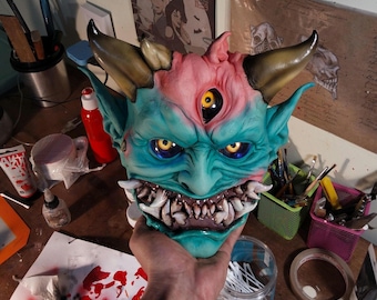 以下が含まれることがあります： 悪魔的なデザインの手描きのマスク。 マスクは、黄色の目、鋭い歯、ピンクとタンの頭を持つターコイズの顔が特徴です。 マスクには3つの目と2つの角があります。 マスクは人の手で持たれています。