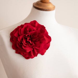 Könnte beinhalten: Eine große, leuchtend rote Stoffblume, die einer Pfingstrose ähnelt, ist an einer weißen Schneiderpuppe befestigt. Die Blütenblätter der Blume sind geschichtet und erzeugen ein volles, strukturiertes Aussehen. Der Hintergrund ist eine weiche, neutrale Farbe.