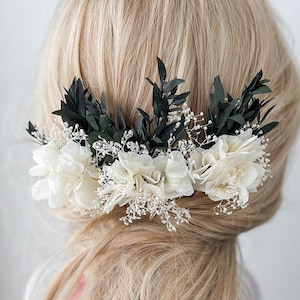 以下が含まれることがあります： 白い花、濃い緑の葉、繊細な白い小花をあしらったヘアコーム。ブロンドの髪にセットされ、柔らかく自然な印象を与えます。結婚式や特別な機会に最適なヘアアクセサリーです。