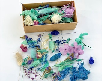 May include: A collection of dried flowers and foliage in shades of blue, green, and purple. The flowers are arranged in a box and include hydrangeas, lavender, and other delicate blooms.