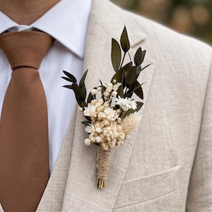 Op de afbeelding: Een corsage met gedroogde crèmekleurige bloemen, kleine witte bloesems en donkergroene bladeren, waarbij de stelen zijn omwikkeld met natuurlijk touw. Het bloemstuk is opgespeld op de revers van een beige linnen colbert.