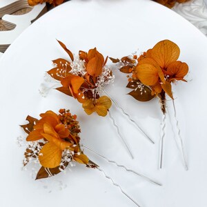 May include: Set of four hair pins with dried orange flowers and white baby's breath. The pins are silver and have a wavy design.