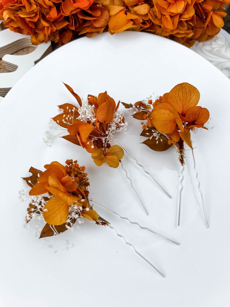 May include: Set of four hair pins with dried orange flowers and white baby's breath. The pins are silver and have a wavy design.